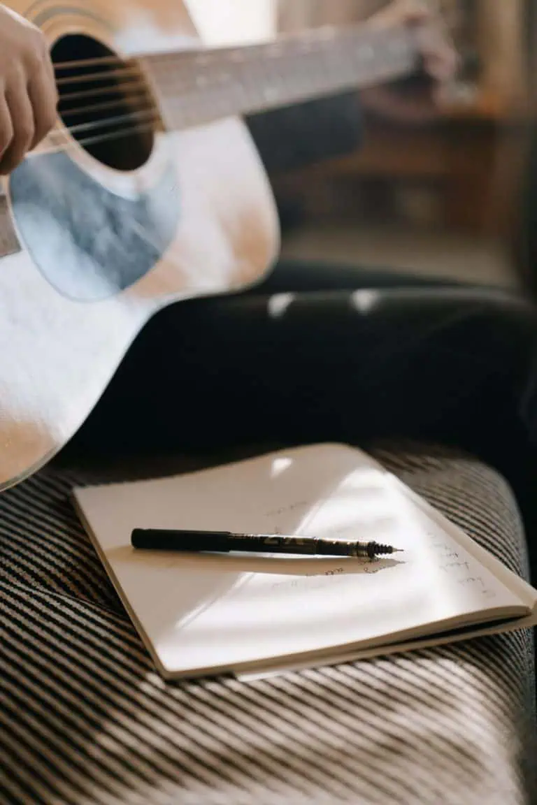 Notebook and pen next to an acoustic guitar while learning how to write a song on guitar